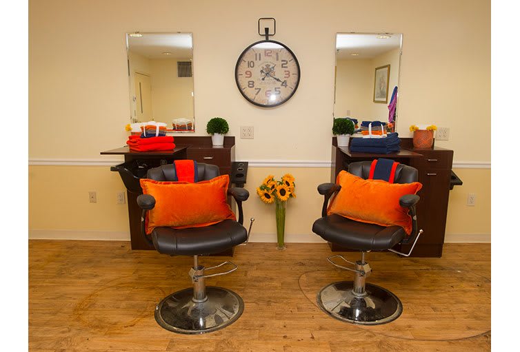 A cozy hair salon with two styling chairs, each adorned with vibrant orange pillows in front of mirrors, and a large clock on the wall.