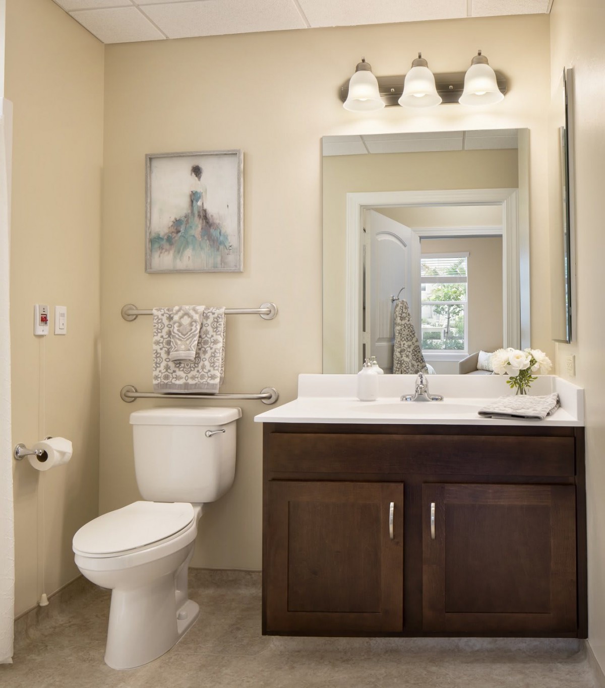 A well-lit bathroom featuring a toilet, sink with a wooden cabinet, large mirror, and decorative artwork on the wall.