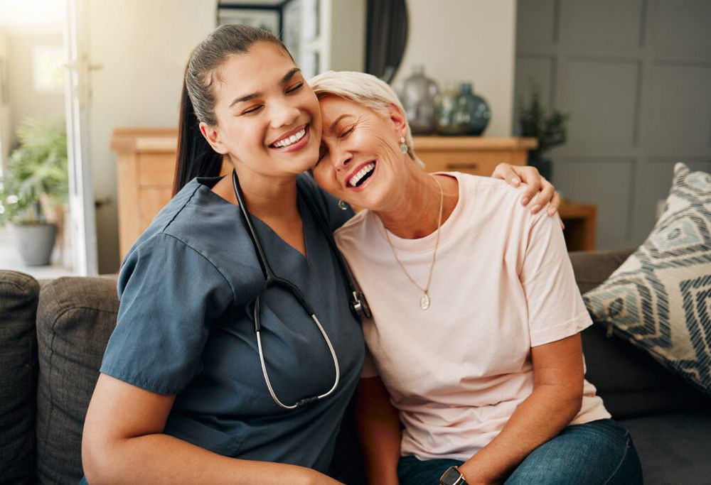 A smiling nurse sitting with a happy woman in a home setting.