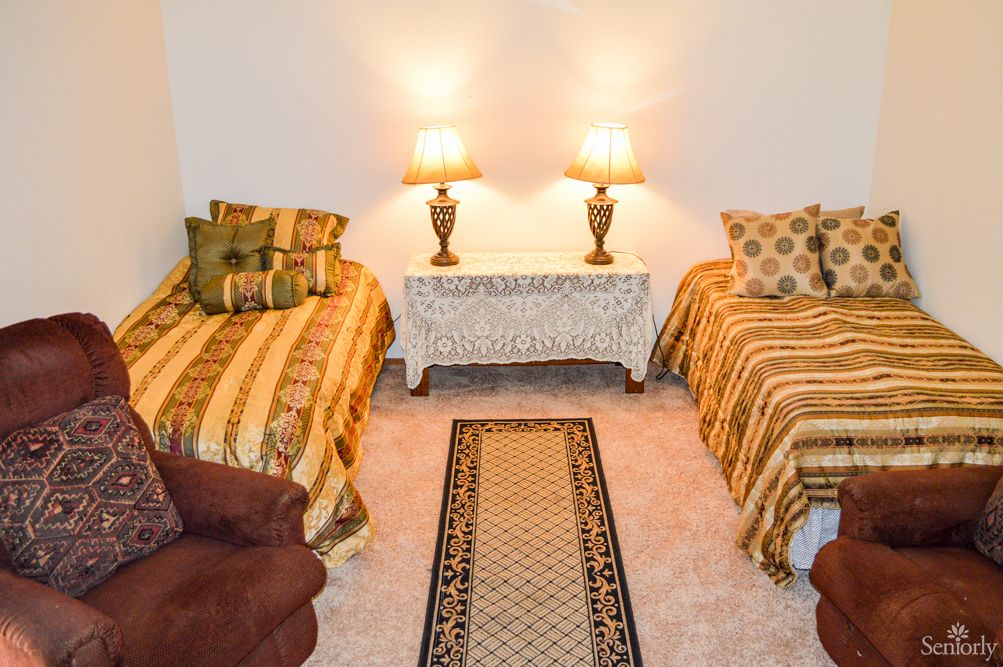 A cozy bedroom featuring twin beds with decorative pillows, a central side table with two lamps, and a patterned carpet on the floor.