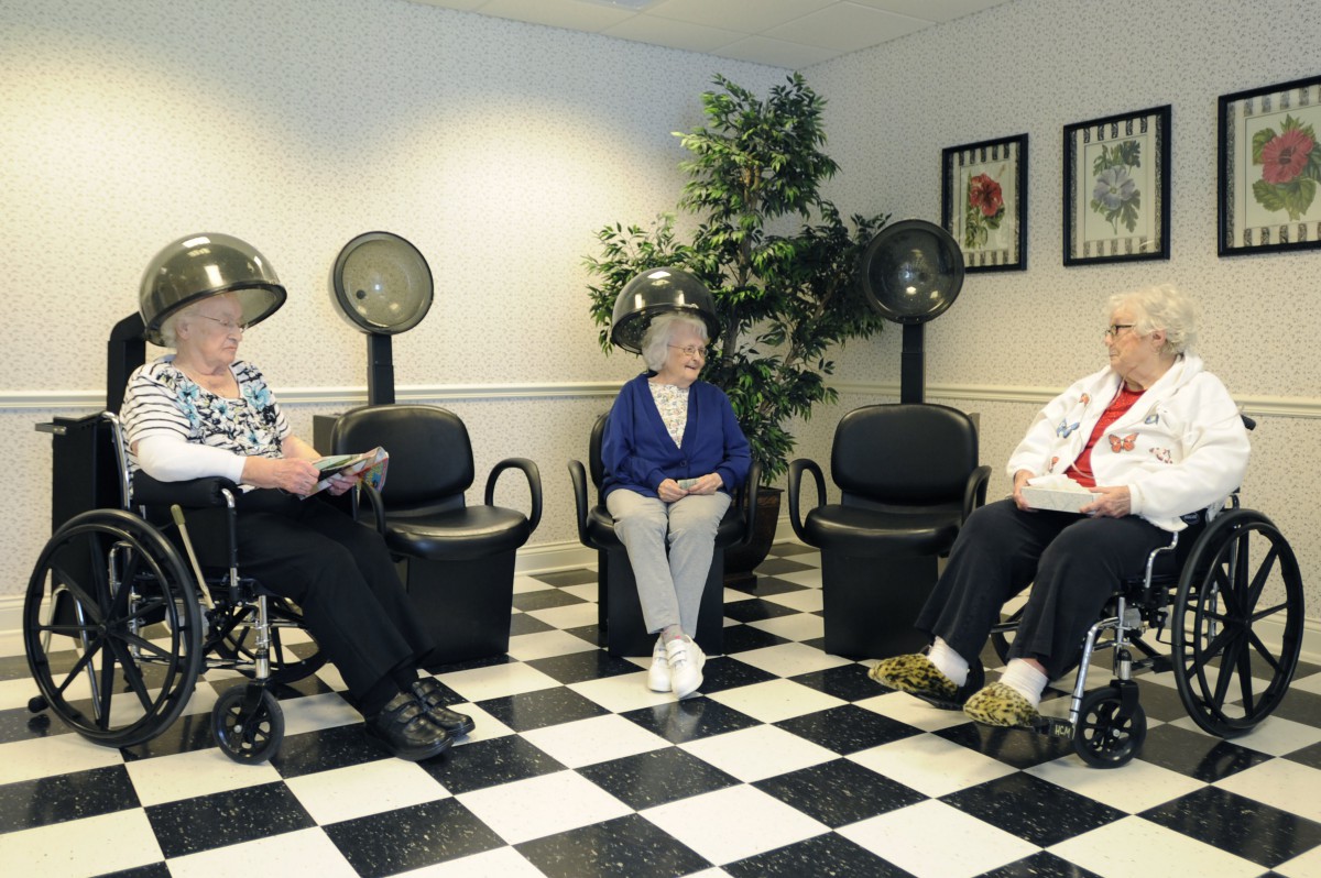Three elderly women sit under hair dryers in a salon with checkered flooring.
