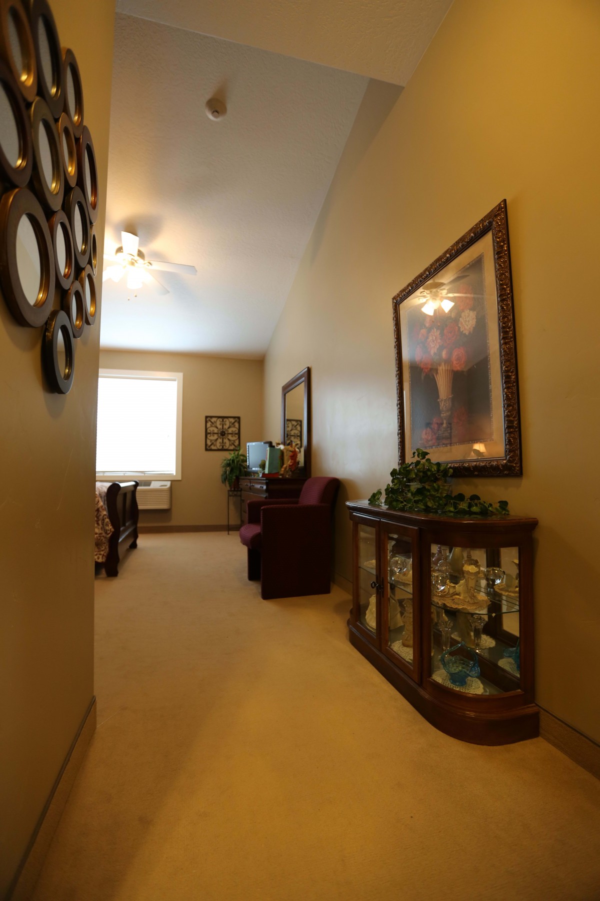 A warmly lit hallway featuring decorative mirrors, a ceiling fan, and elegant furniture with a glass display cabinet.