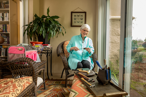 An elderly woman engaged in a crafting activity in a cozy indoor space, surrounded by sunlight and plants.