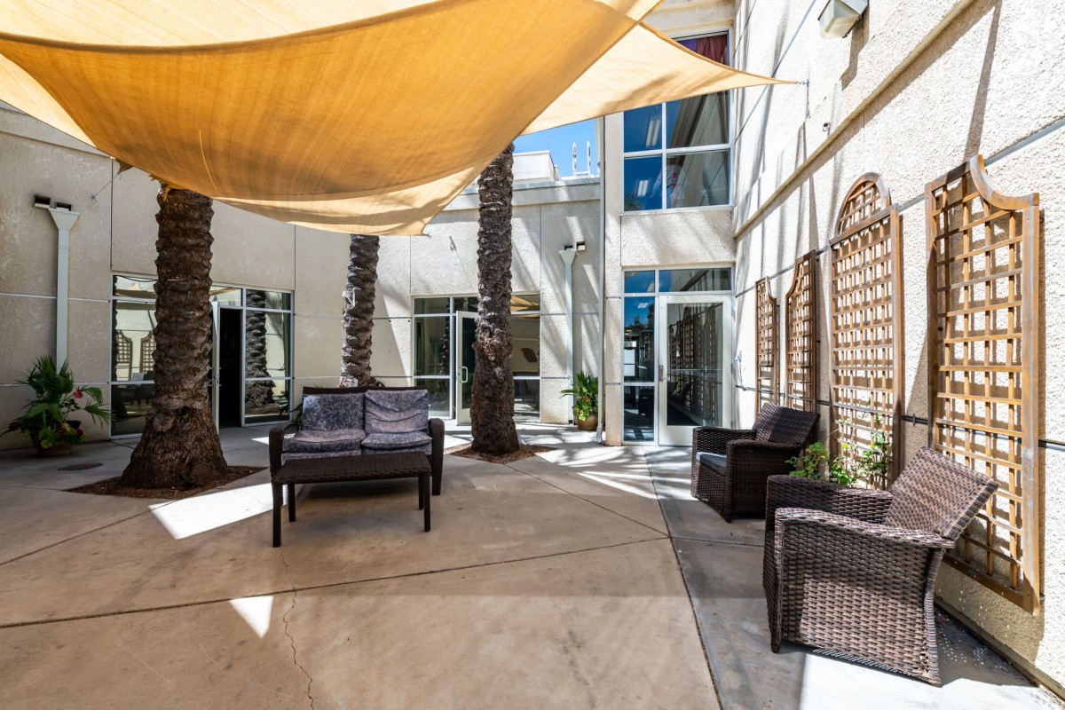 A sunny outdoor patio with shade sails, wicker furniture, and palm trees.