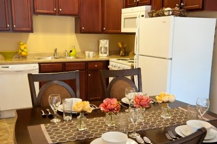 A cozy kitchen with a dining table set elegantly for a meal, featuring floral decorations and kitchen appliances in the background.