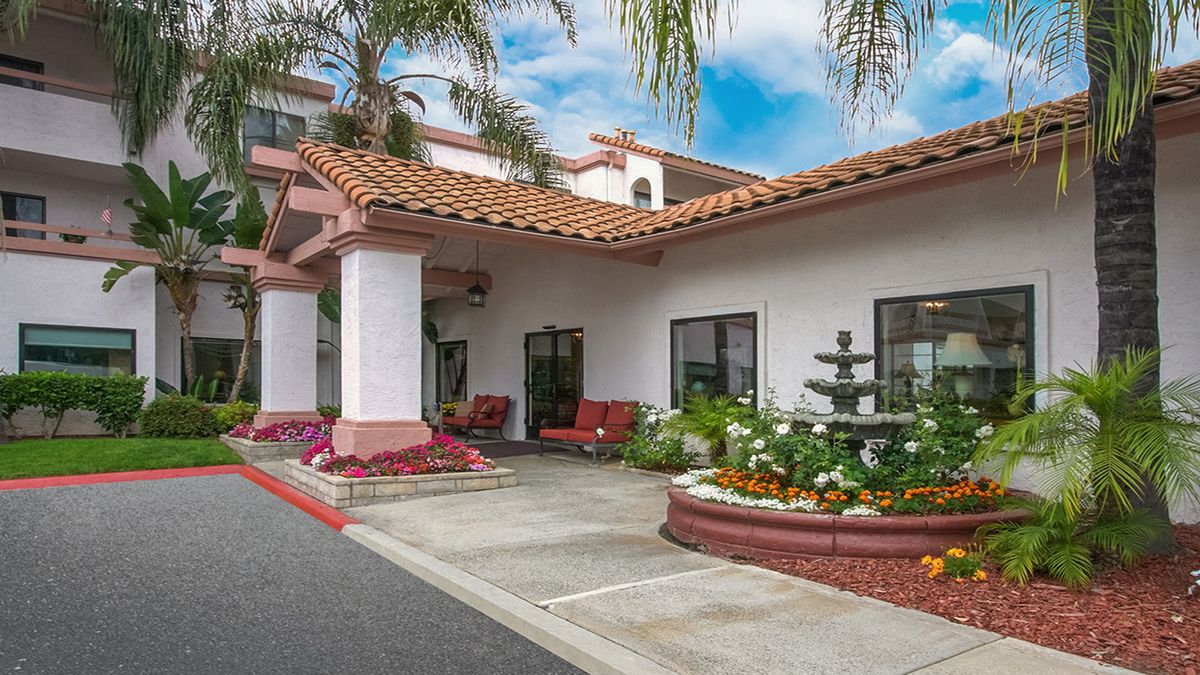 The image shows the entrance of a hotel with landscaped gardens and outdoor seating.