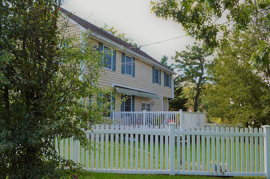 A suburban house with a white fence and surrounding trees.