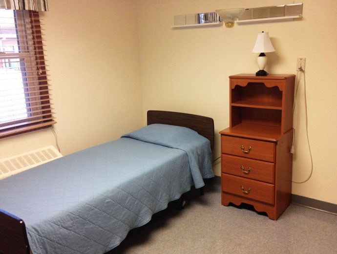 A simple, minimalist bedroom with a single bed, dresser, and lamp.