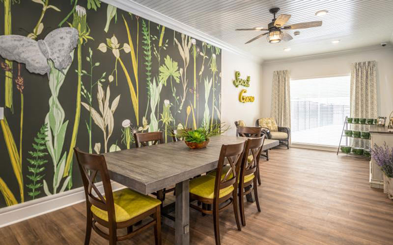 A modern dining room featuring a large wooden table, surrounded by yellow-cushioned chairs. The room is adorned with a vibrant floral wallpaper, adding a touch of nature to the contemporary interior.