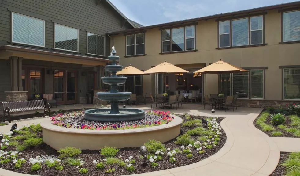 A courtyard featuring a central fountain surrounded by flowers, with outdoor seating shaded by umbrellas in front of a two-story building.