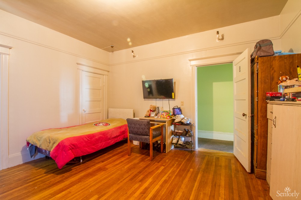 A cozy bedroom featuring a single bed, desk, and television on the wall.