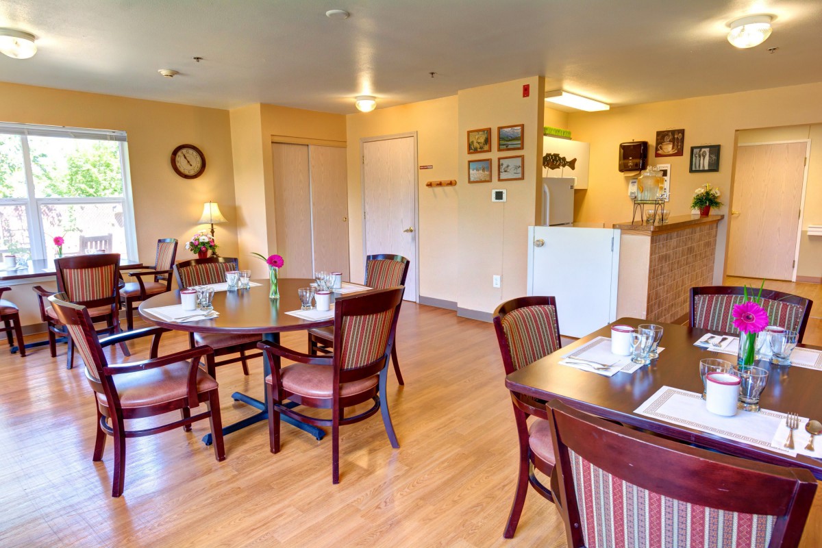 A cozy dining area with neatly arranged tables and chairs, featuring a kitchen space in the background.