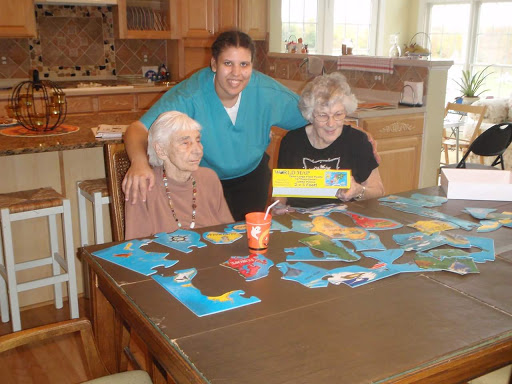 A caregiver enjoys a leisurely moment with two elderly women while playing a board game in a cozy kitchen setting.