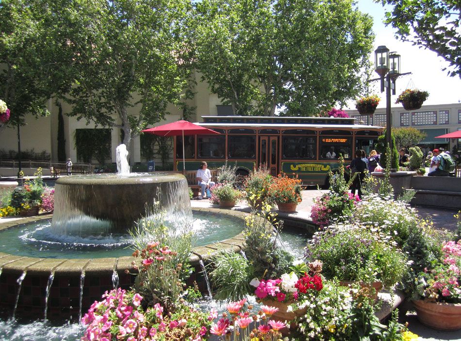 A scenic view of a park with a fountain, colorful flowers, and a trolley in the background.