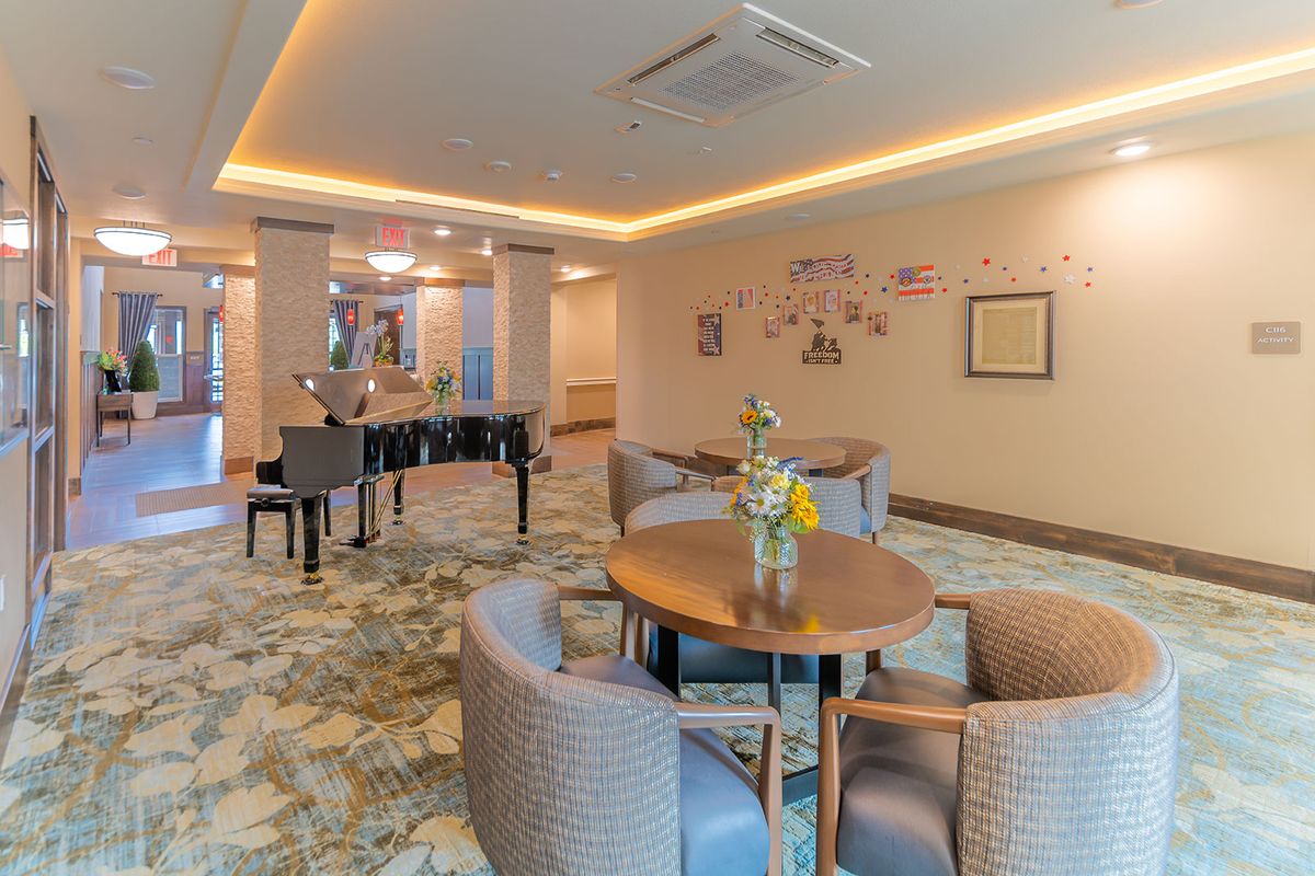 A cozy lounge area with a piano, round tables, and comfortable chairs, featuring decorative lighting and carpeted flooring.