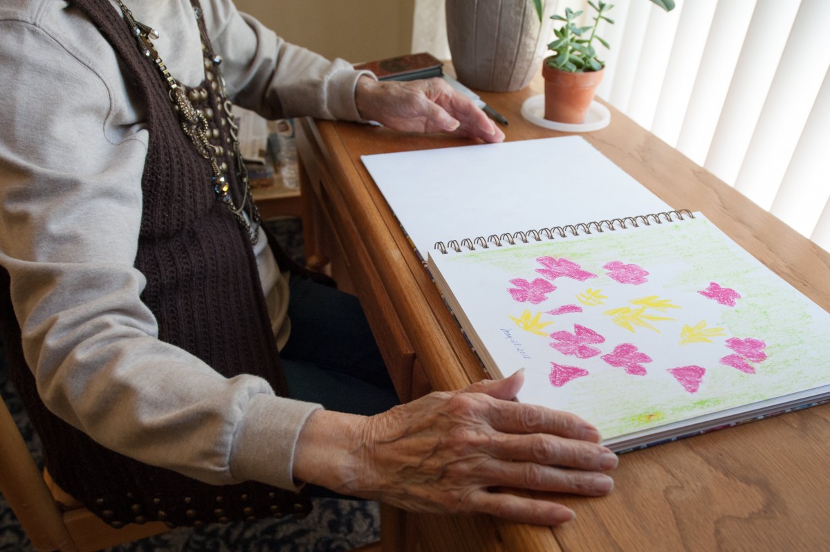 An elderly person sitting at a table, working on a colorful drawing in a sketchbook.
