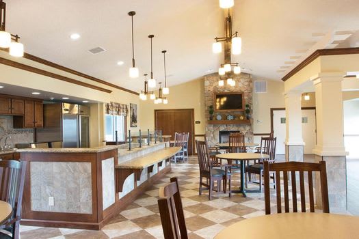 A cozy restaurant dining area featuring wooden tables and chairs, elegant lighting fixtures, a checkered floor, and a central stone fireplace.