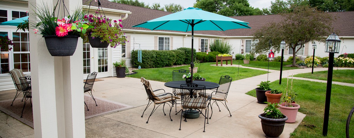 A cozy backyard patio with a table and chairs set under a teal umbrella, surrounded by lush greenery.