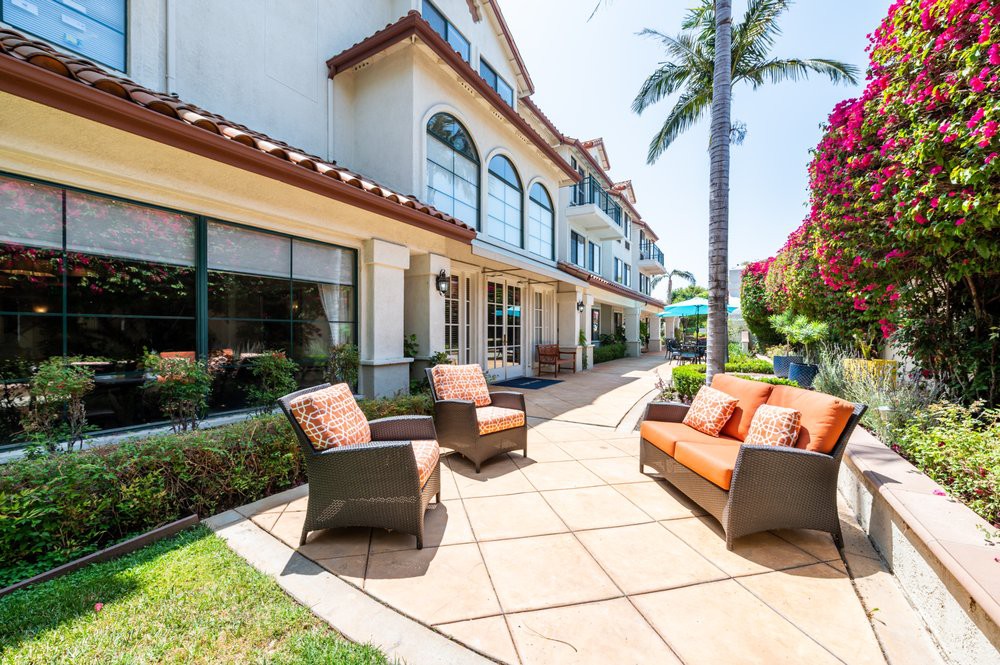 A sunny outdoor patio area with comfortable furniture in front of a multi-story building, featuring a palm tree and well-maintained landscaping.