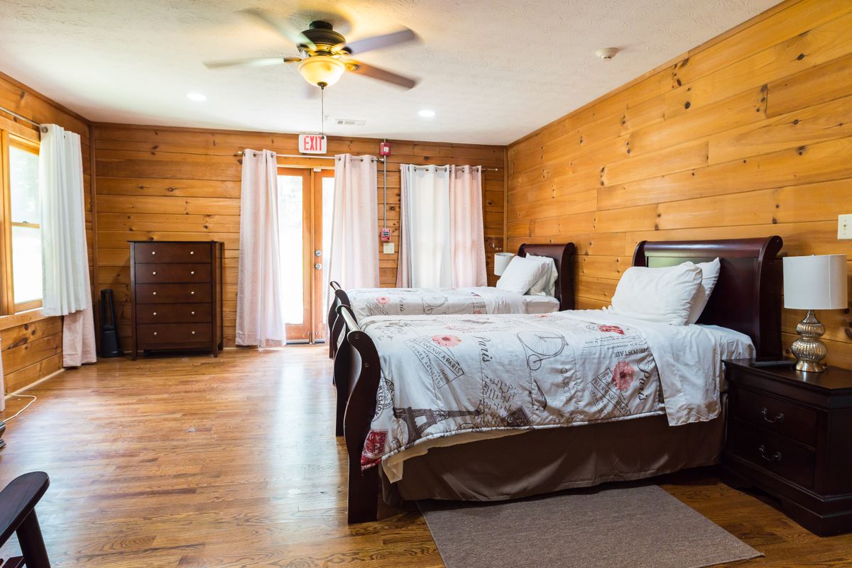 A cozy bedroom with wooden walls featuring two beds, a ceiling fan, and a dresser.