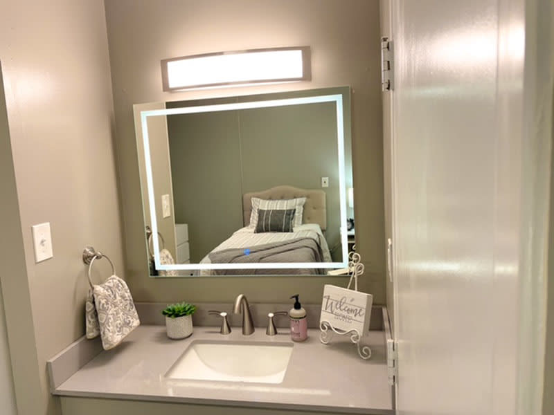 A modern bathroom vanity with a lighted mirror, reflecting a neatly made bed in an adjoining room.