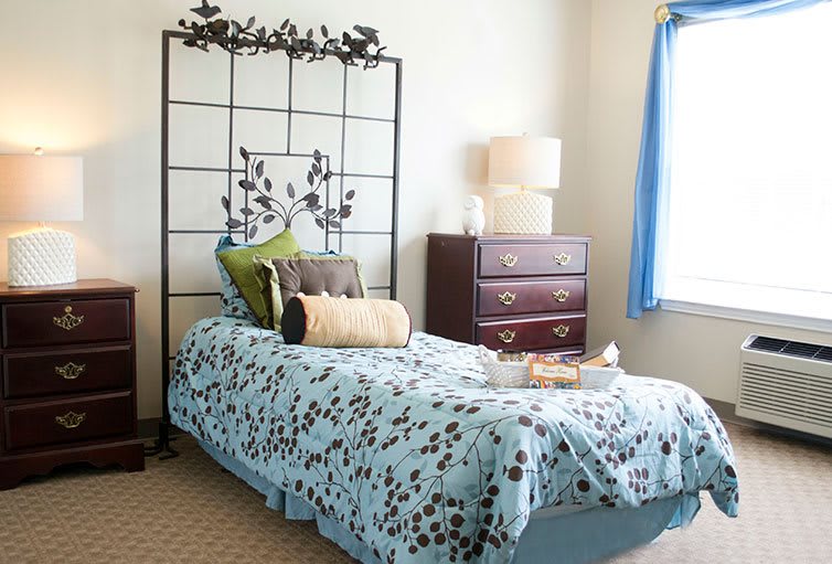 A cozy bedroom with a decorative headboard, patterned bedding, and a bedside lamp on a dresser.