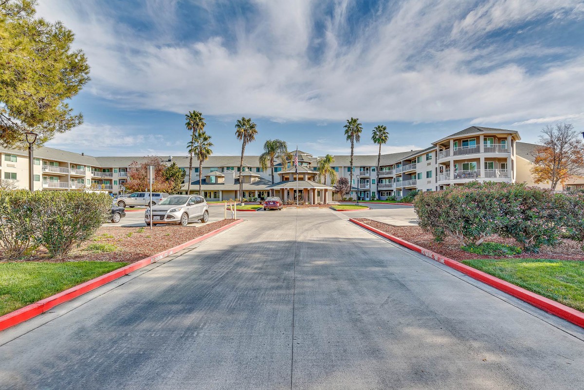A building with palm trees, a parking lot, and a road leading to its entrance under a cloudy sky.