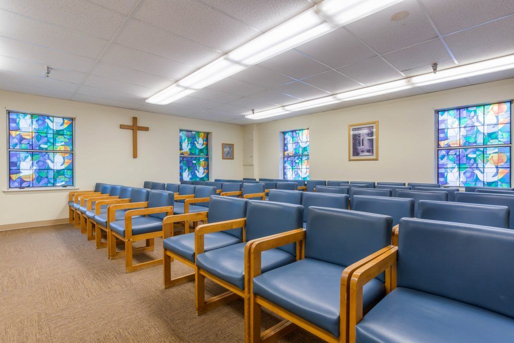 An interior view of a chapel with rows of blue cushioned chairs and a wooden cross on the wall.