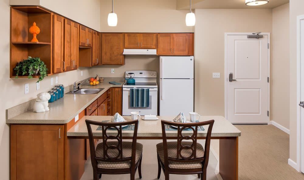 A modern kitchen with wooden cabinets, featuring a stove, refrigerator, and a small dining area with two chairs.