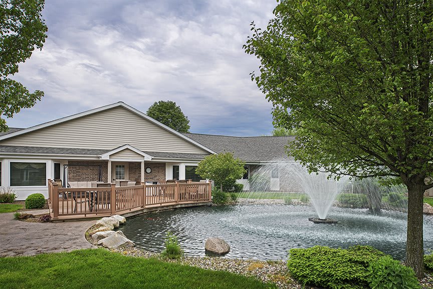 A scenic view of a building with a wooden bridge over a pond featuring a fountain, surrounded by trees and under a partly cloudy sky.