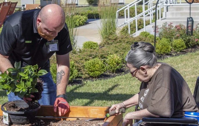 A volunteer assists an elderly person with gardening in a community garden.