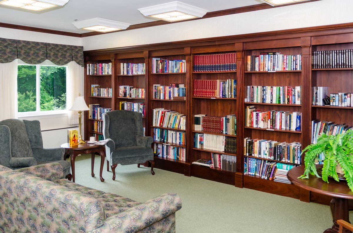 A cozy reading room with bookshelves full of books, a comfortable chair, and a small table with a lamp.