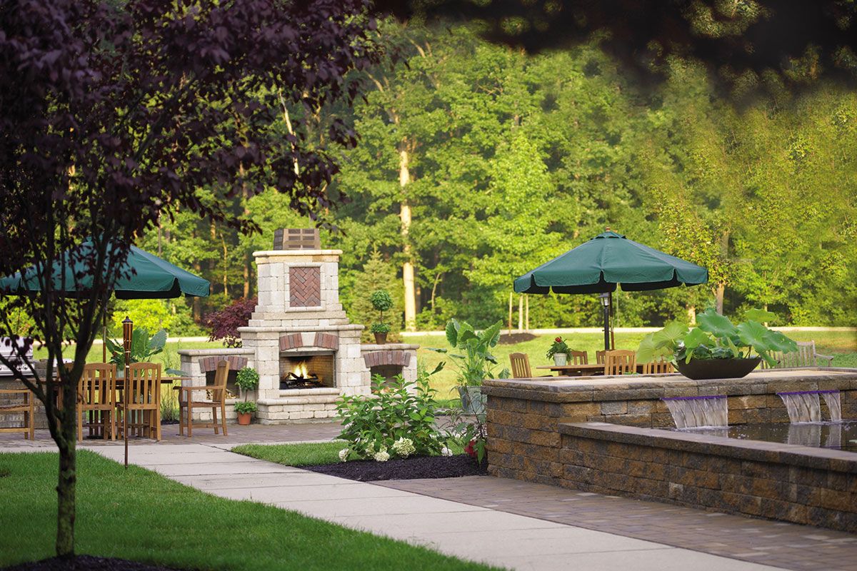 A serene garden patio with a stone fireplace, surrounded by lush greenery and shaded seating areas.