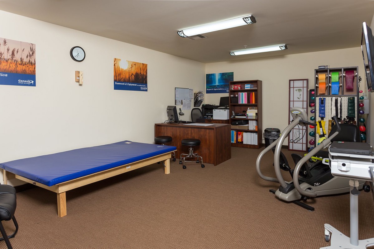 A room equipped with exercise machines, a desk, bookshelves, and a therapy table.