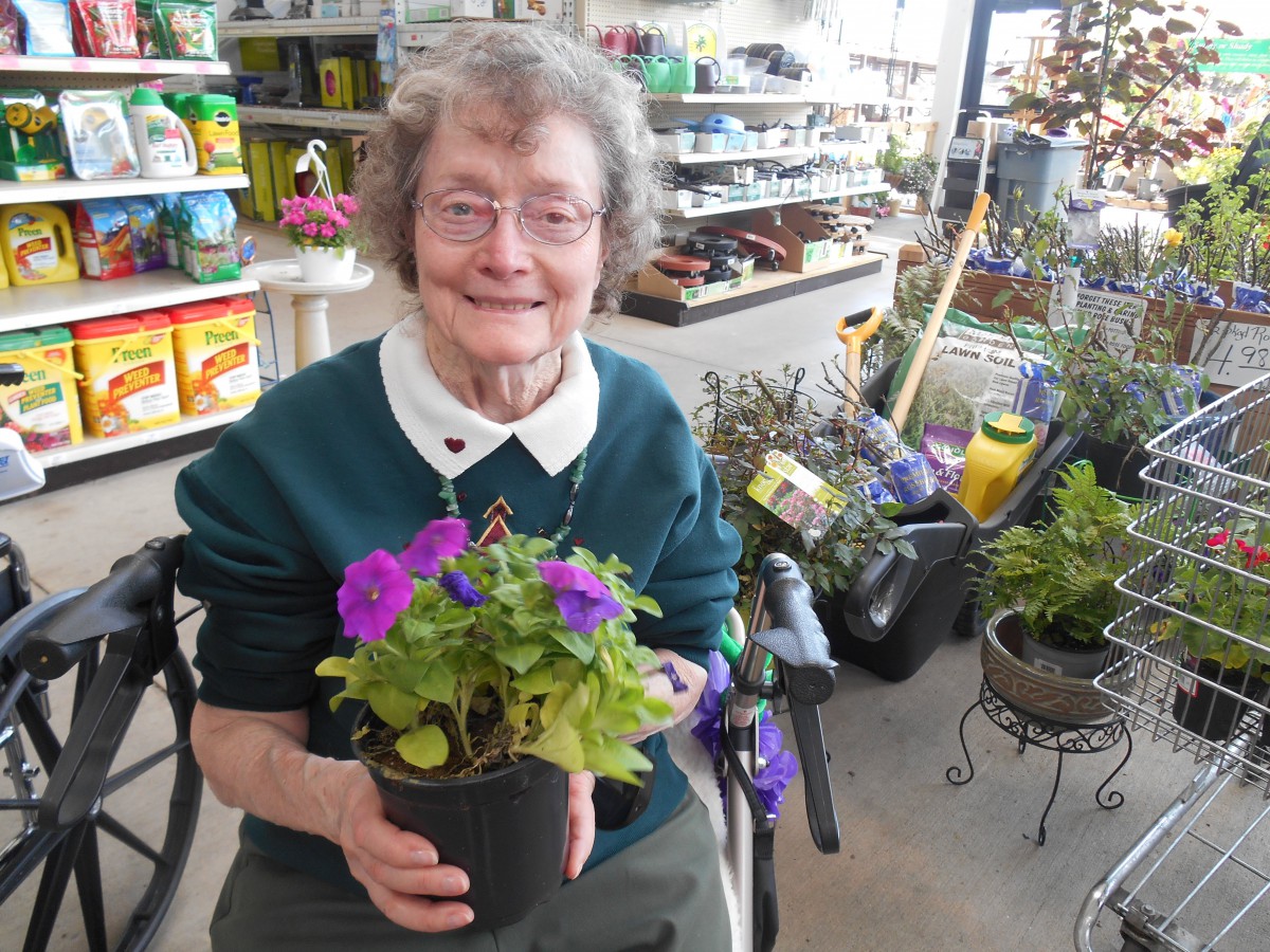 An elderly woman smiling while holding a potted plant with purple flowers at a garden center.