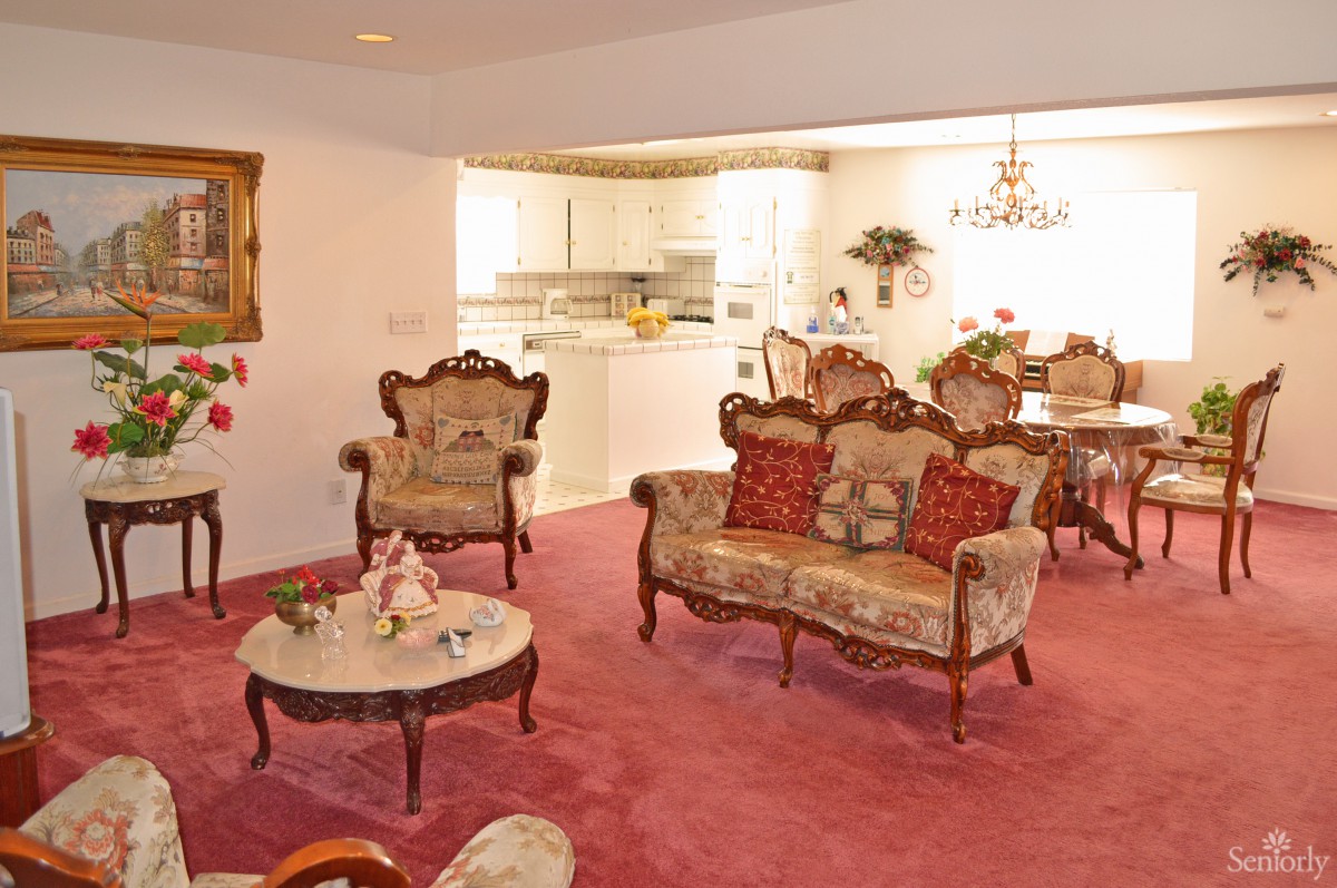 A cozy living room with vintage-style furniture and an open kitchen, featuring a red carpet and elegant chandelier.