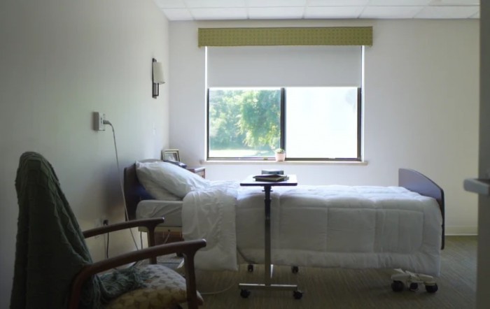 Empty hospital room with a neatly made bed and a view of greenery through the window.