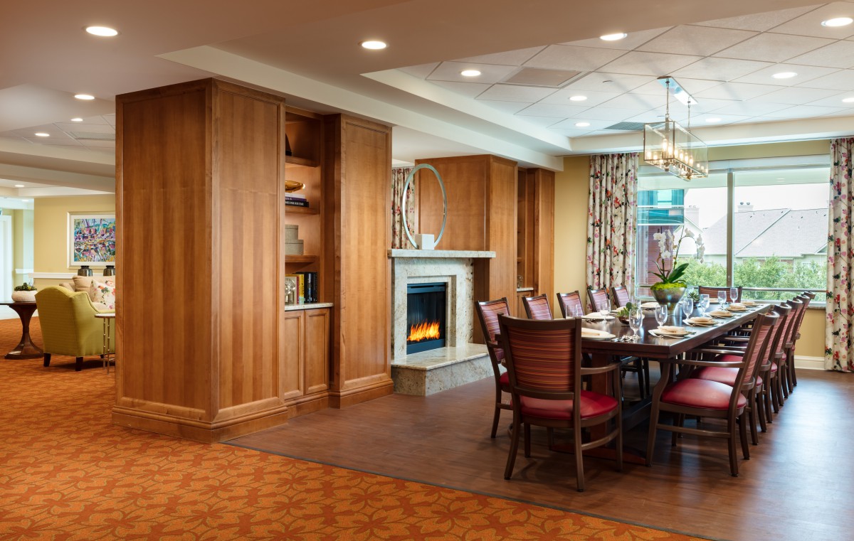 A warmly decorated dining room featuring a wooden table surrounded by chairs, with a fireplace and elegant chandelier.
