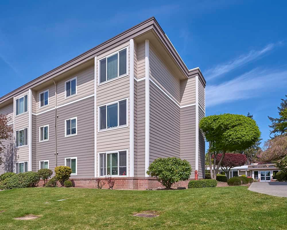 A modern, multi-story apartment building with well-maintained landscaping and a clear blue sky in the background.