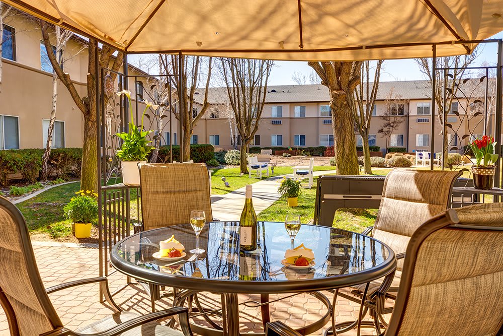 A cozy outdoor patio setup with a table and chairs under an umbrella, featuring wine glasses and a bottle, overlooking a well-maintained garden in an apartment complex.