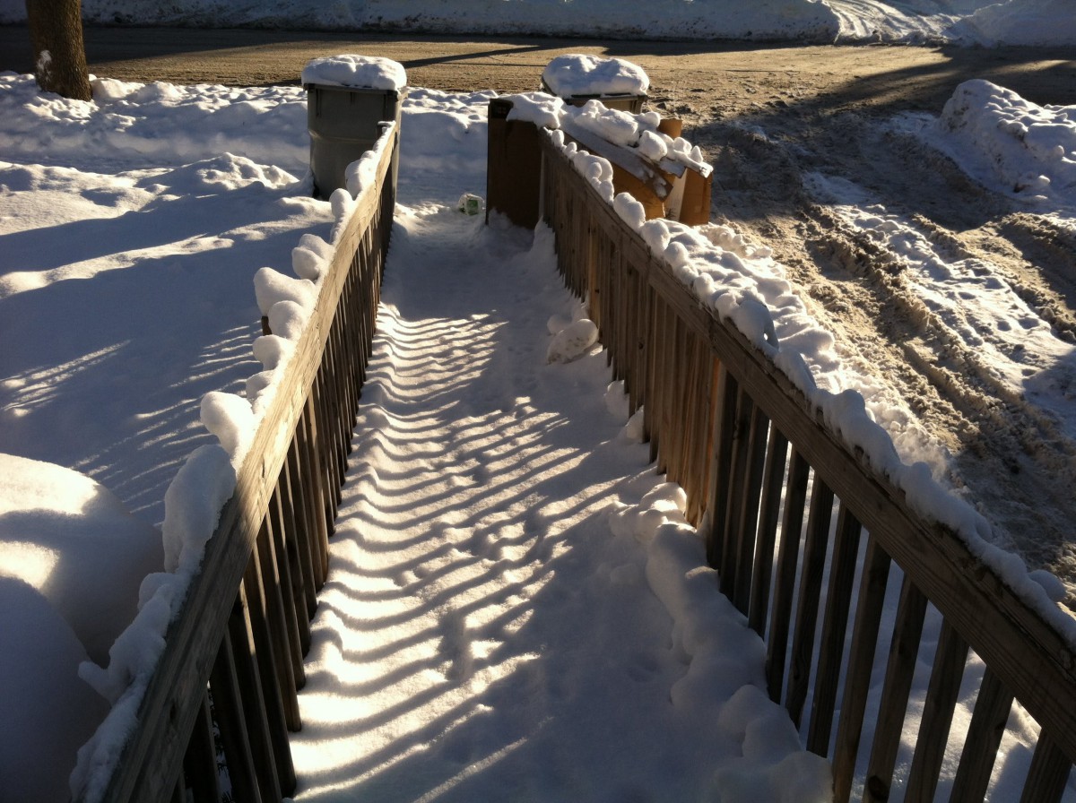 A snow-covered pathway lined with wooden fences casting shadows in the winter sunlight.