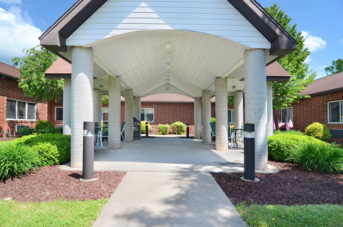 A covered walkway with columns leading to a building entrance, surrounded by landscaped greenery.