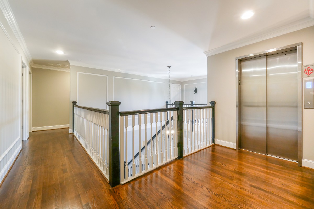 A well-lit hallway with hardwood flooring, featuring a staircase with a dark banister and an elevator.