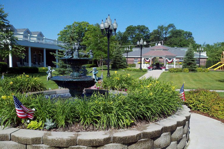 A beautiful garden with a central fountain, surrounded by lush greenery and a pathway leading to a decorative building.