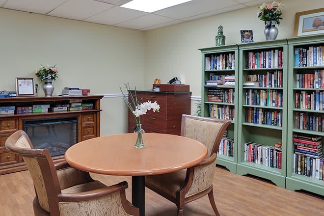 A cozy office space featuring a round table with two chairs, a bookshelf filled with books, and a vase with flowers on the table.
