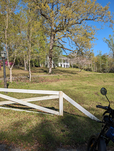 A scenic view of a rural landscape featuring a white gate, a motorcycle, and a house nestled among trees under a clear blue sky.