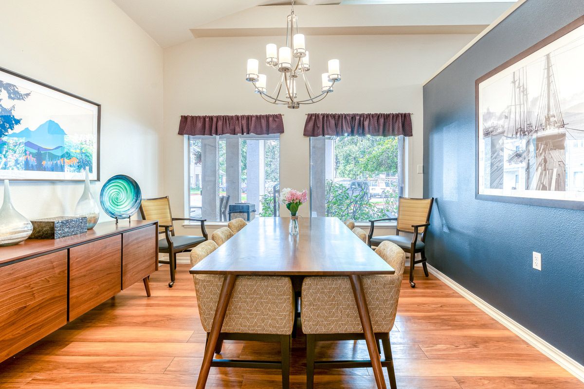 A bright and elegant dining room featuring a long wooden table, chairs, and a chandelier, with large windows letting in natural light.