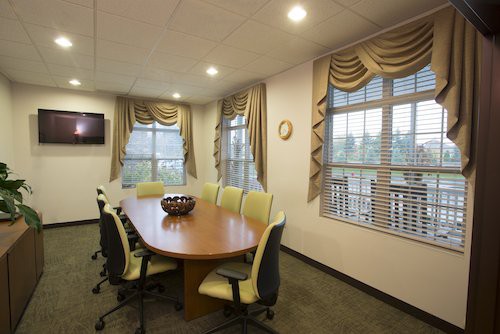 A well-lit conference room with a long table and chairs, featuring large windows with curtains.