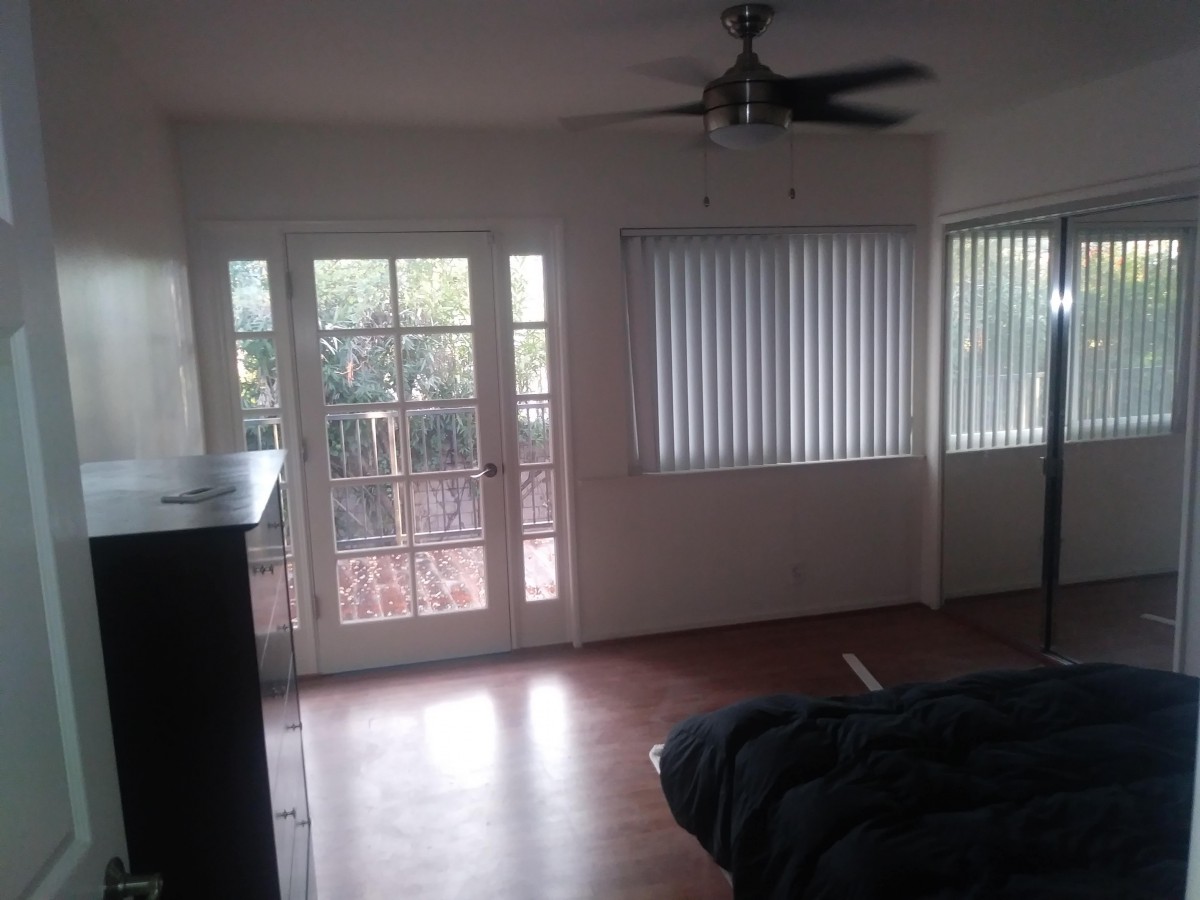 A cozy living room featuring a ceiling fan, large windows with blinds, and a door leading to an outdoor area.