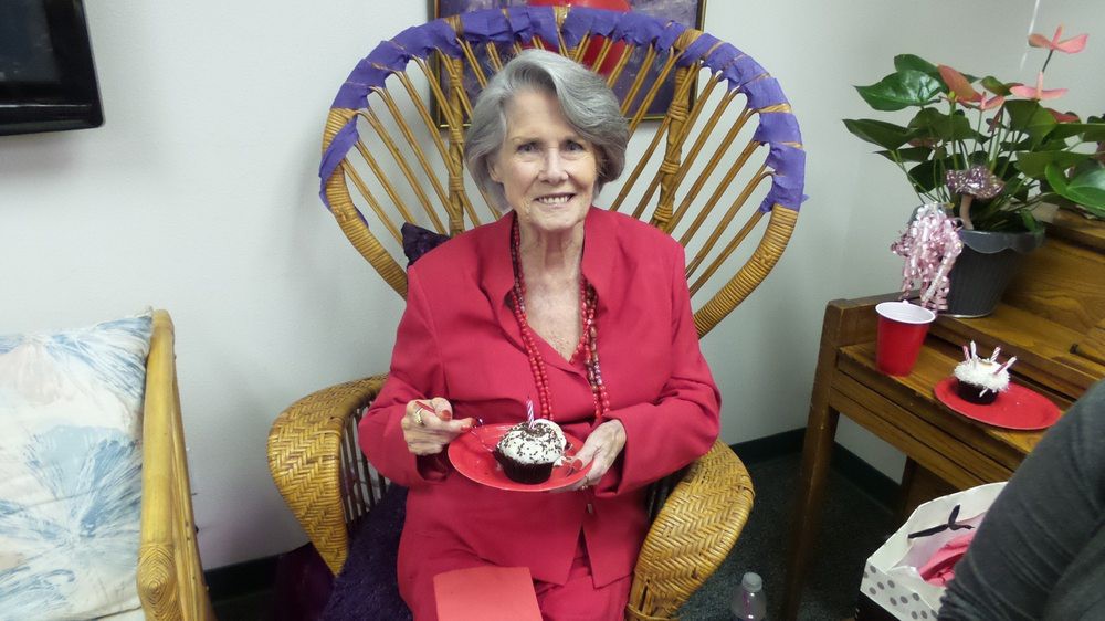 An elderly woman in a red outfit is smiling while holding a cupcake, seated indoors on a decorative chair.